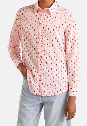 Personne portant une chemise rose clair à boutons avec un motif losange rouge et un jean bleu clair, une main dans la poche, sur fond blanc.