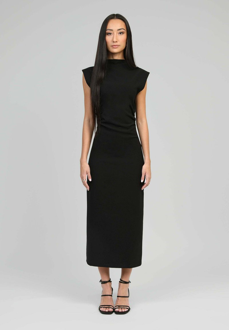 Robe noire ajustée avec des manches courtes en forme de cap, un col haut et une longueur au genou. Texture lisse et design minimaliste, complétée par des talons à lanières.