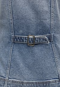 Gros plan sur un tissu en denim bleu avec une sangle horizontale fixée par une boucle métallique rectangulaire.