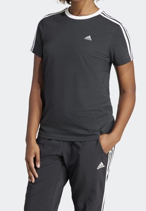 Personne portant une tenue de sport Adidas noire avec des rayures blanches sur les manches et le pantalon, debout avec une main dans la poche.