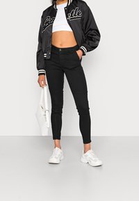 Sort satin bomberjakke med hvid "Seattle" tekst, cropped hvid top, sorte skinny jeans, hvide sneakers og en hvid håndtaske.