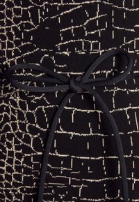 Tissu noir avec un motif abstrait texturé en lignes beiges. Présente un nœud papillon noir, soulignant une taille bien marquée.