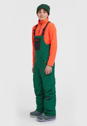 Groene sneeuwoveralls met zwarte accenten en verstelbare banden, gecombineerd met een oranje langemouwen top en grijze winterlaarzen.