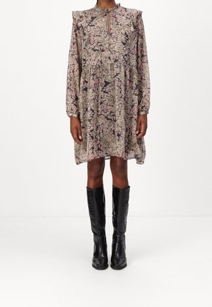 Kobieta ubrana w luźną, długorękawową sukienkę sięgającą do kolan z przytłumionym abstrakcyjnym wzorem i czarne, skórzane kozaki do kolan stoi na białym tle.