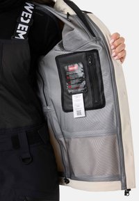 Veste imperméable aux couleurs noire et beige. Elle dispose d'une poche en mesh, d'une étiquette avec la technologie RECCO et de détails réglables en matière de quincaillerie.