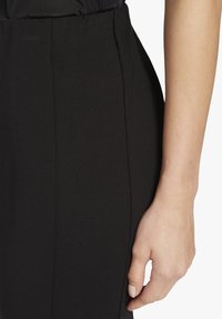 Zwarte aansluitende rok met een gladde textuur en zijnaad. Een hand is nabij de tailleband gepositioneerd, wat een natuurlijke huidtint laat zien.