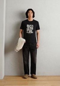 T-shirt nera con stampa grafica bianca, jeans scuri, scarpe beige e un maglione beige tenuto nella mano sinistra, in piedi su un pavimento in legno.