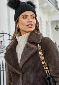 Abrigo de piel sintética marrón con cuello de chal, sobre un suéter de punto trenzado color crema. La modelo lleva un gorro negro con pompones y pendientes de aro plateados.