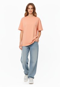 T-shirt oversize pesca con maniche corte e scollatura rotonda, abbinata a jeans larghi blu chiaro e sneakers bianche.