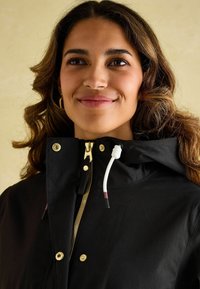 Czarna kurtka z kapturem, z złotym zamkiem i elementami, wyposażona w biały sznurek do regulacji oraz widoczne szwy na fakturowanej tkaninie.