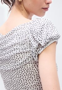 Λευκό ύφασμα με μαύρες πουά, με φουσκωτά μανίκια και ρυτιδωτή υφή στο πίσω μέρος. Ελαφρύ, διαφανές υλικό.