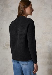 Schwarzer, gerippter Strickpullover mit langen Ärmeln und rundem Ausschnitt, kombiniert mit hellblauen Jeans, vor einem strukturierten neutralen Hintergrund.