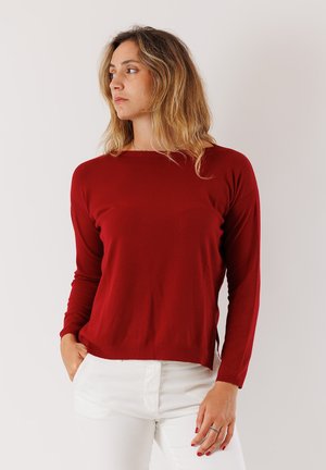 Donna con capelli biondi alla spalla, che indossa un maglione rosso a maniche lunghe e pantaloni bianchi, mentre guarda verso sinistra con una mano in tasca.