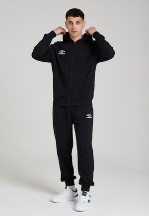 Felpa nera con zip e tasca a marsupio, con logo. Abbinata a pantaloni jogger neri coordinati. Scarpe da ginnastica bianche con accenti verdi.