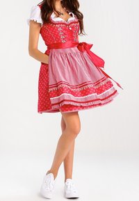 Robe dirndl rouge avec accents blancs, encolure en dentelle, corsage à motifs, jupe plissée et un grand nœud en satin à la taille. Associée à des baskets blanches.