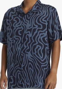 Camicia a maniche corte di colore blu navy con motivi ondulati azzurri. Realizzata in tessuto morbido con chiusura a bottoni e vestibilità rilassata.