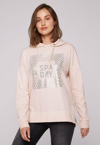 Felpa rosa chiaro con una grafica argento che riporta la scritta "SPA DAY" in un design testurizzato. Presenta un cappuccio regolabile con coulisse e fessure laterali all'orlo.
