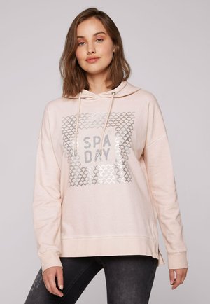 Sudadera rosa claro con un gráfico plateado que dice "DÍA DE SPA" en un diseño texturizado. Cuenta con capucha con cordón y slits laterales en el dobladillo.