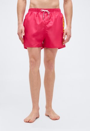 Homme adulte portant un short de bain rouge avec du texte jaune sur le côté, debout pieds nus sur un fond blanc uni.