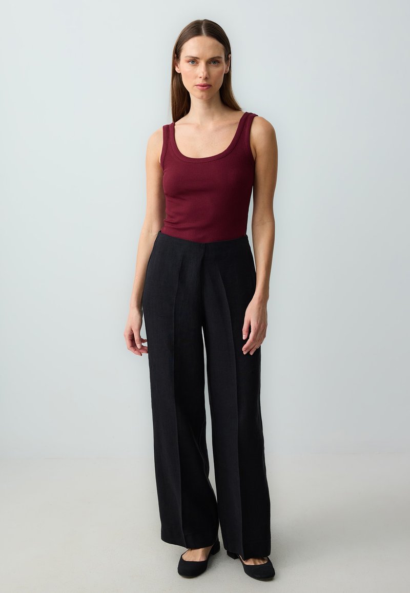 Femme debout sur un fond neutre, portant un haut sans manches rouge foncé, un pantalon noir à jambes larges, et des chaussures noires.