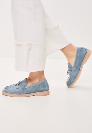 Persona que lleva mocasines de ante azul claro con suelas beige y detalle de borlas, combinados con pantalones blancos recortados con dobladillo deshilachado.