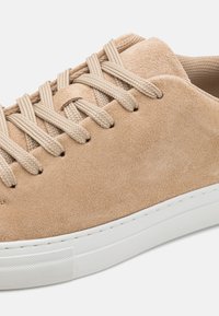 Kasta zamšveida sneakers ar auzu krāsas saitēm, baltu gumijas zoli un teksturētu virsmu. Iespējo minimālisma dizains un tīras līnijas.