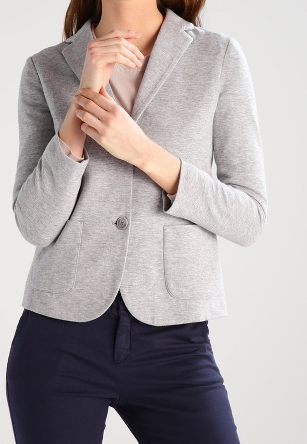 Femme portant un blazer gris clair boutonné une fois sur un top beige, avec un pantalon bleu foncé, ajustant ses mains près de sa poitrine.