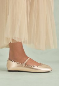 Ballerines métalliques dorées avec bords festonnés et une bride, présentant une texture lisse et un bout pointu. Portées avec une jupe légère en tulle transparent.