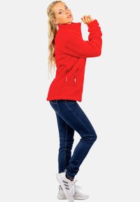 Roter Fleece-Pullover mit hohem Kragen, seitlichen Reißverschlusstaschen und strukturiertem Stoff. Kombiniert mit enganliegenden blauen Jeans und weißen Sportschuhen.