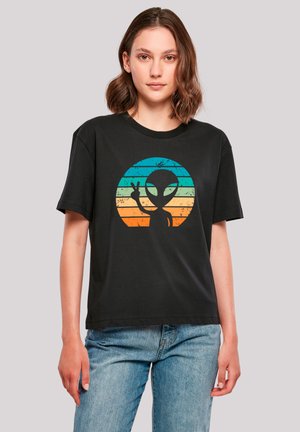 Young woman wearing black t-shirt with colorful retro-striped alien graphic making peace sign, paired with blue jeans, standing against plain background.