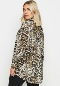 Koszula w cętki z lamparta o jedwabistej fakturze, z długimi rękawami i zaokrąglonym dołem. Paleta kolorów obejmuje odcienie brązu, czerni oraz subtelne złote akcenty.