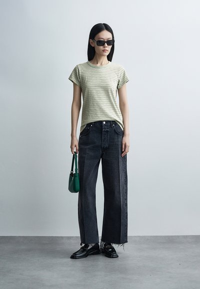 Γυναίκα που φοράει γυαλιά ηλίου, ανοιχτοπράσινο ριγέ μπλουζάκι, φαρδιά μαύρα τζιν, μαύρα παπούτσια και κρατάει μια μικρή πράσινη τσάντα χειρός σε απλό φόντο.