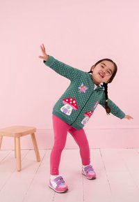 Zelená s bodkami bunda s nášivkami húb a víl, ružové legíny a farebné tenisky. Pozadie je svetloružové.