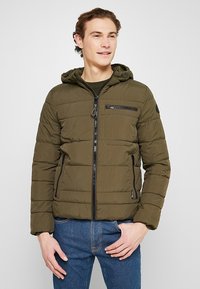 Olijfgroene pufferjack met capuchon, ritssluiting aan de voorkant en borstzak, ribbels aan de mouwen en een gestructureerde afwerking. Zwarte accenten op de ritsen.