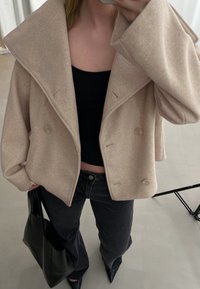 Beige wollen blend cropped jasje met grote kraag, dubbele knopen en losse mouwen; gecombineerd met een zwarte top en zwarte jeans.