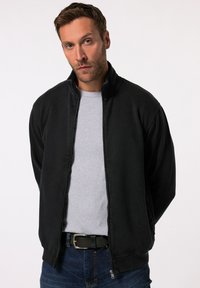 Chaqueta negra con cremallera, cuello y puños acanalados, que presenta una textura suave y cremallera completa, llevada sobre una camiseta gris clara.
