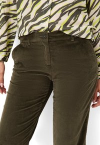 Olivgrüne Cord-Hosen mit vertikaler Rippenstruktur, ausgestattet mit einem Knopf- und Reißverschluss. Kombiniert mit einer gemusterten Bluse.