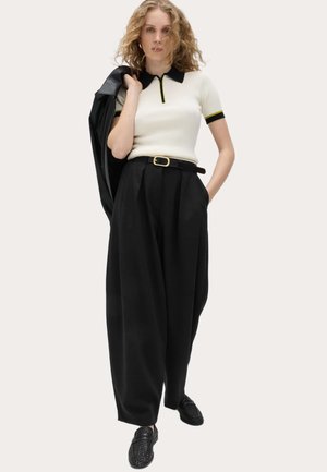 Frau mit lockigem Haar, die ein weißes Kurzarm-Poloshirt, schwarze Hose mit weitem Bein und Gürtel, eine schwarze Jacke über der Schulter und gewebte Loafers trägt.