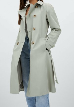 Femme portant un trench-coat gris clair croisé avec ceinture et un jean bleu, debout avec une main dans la poche du manteau.