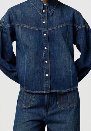 Mørkeblå denimjakke med hvide trykknapper kombineret med matchende jeans med frontlommer og kontrastsyninger.