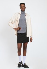 Camicia oversize beige con dettagli a borchie, dolcevita grigio, gonna nera aderente, calze bianche fino al ginocchio e mocassini neri con una catena dorata.
