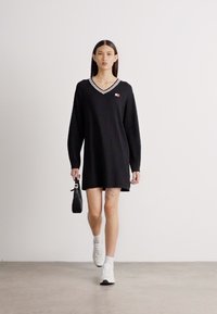 Μαύρο φόρεμα με V γιακά και μακριά μανίκια, λευκό ριγέ γιακά και μία μικρή ετικέτα λογότυπου. Το μοντέλο φορά λευκά αθλητικά και κρατά μία μίνι τσάντα.