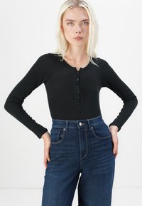 Crna pamučna majica dugih rukava s rebrastim uzorkom i detaljima dugmadi, u kombinaciji s plavim trapericama visokog struka koje imaju klasičan prednji zatvarač i džepove.