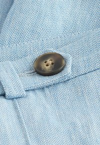 Tissu bleu clair avec un motif en chevrons, comportant un bouton marron à quatre trous, fixé avec une languette en tissu.