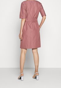 Růžové šaty s krátkým rukávem, se zúženým pasem, délka po kolena a hladká textura. Nalezené s bílými sandály na vysokém podpatku s pruhovanými detaily.