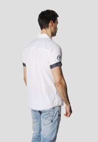 White short-sleeve shirt with dark blue accents on sleeves, featuring a logo patch on the left arm and a classic collar design.