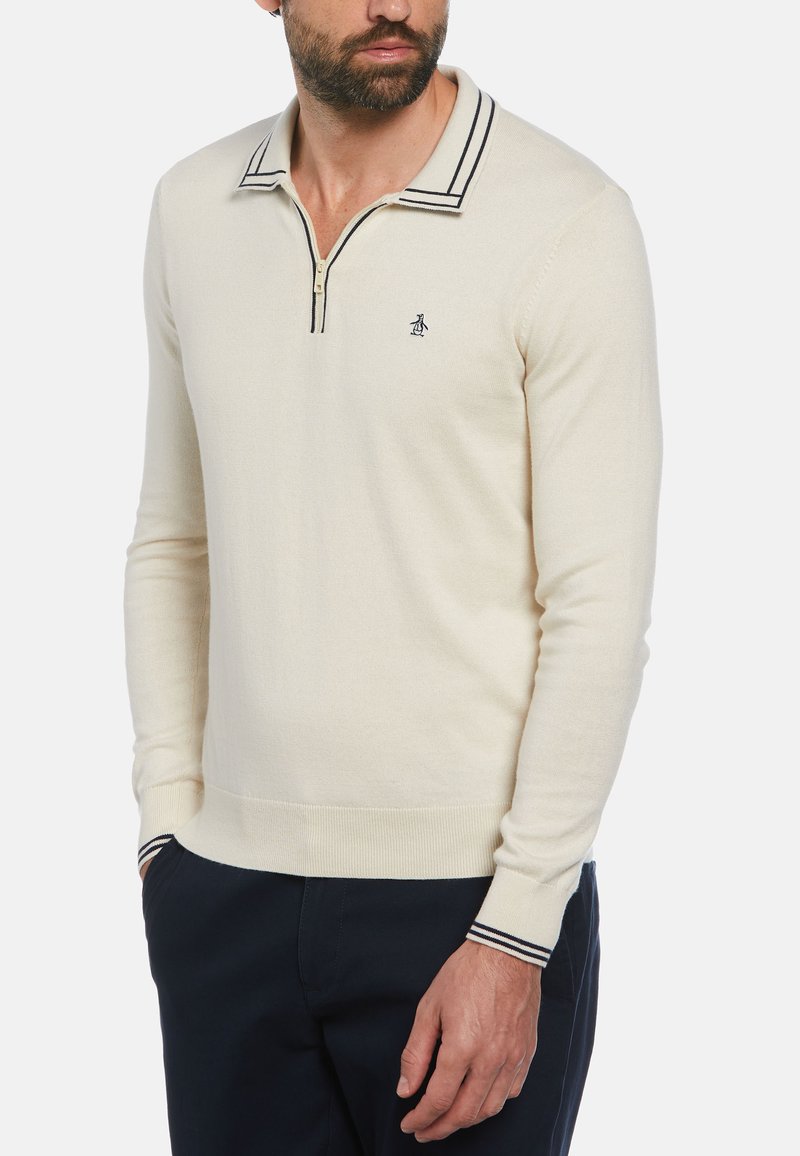 Pull-over en tricot beige avec un col zippé, featuring des accents rayés bleu marine sur le col et les poignets. Petit logo sur la poitrine.