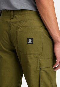 Pantaloni cargo verde oliva con tessuto testurizzato, dotati di una tasca posteriore e un'etichetta nera Timberland, che mostrano cuciture dettagliate.