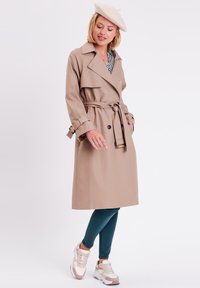 Abrigo trench beige con cintura ajustada, botón frontal y cuello. Llevado sobre una blusa estampada, combinado con leggings en color verde azulado y zapatillas beige.