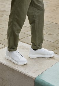 En person med vita sneakers och olivgröna byxor står på en betongkant med en stenlagd mark i bakgrunden.